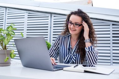 Başarılı genç bayan girişimci gülümseyerek bir bilgisayar ekranına bakıyor, açık kafe terasında dizüstü bilgisayar kullanarak çalışıyor.