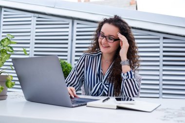 Başarılı genç bayan girişimci gülümseyerek bir bilgisayar ekranına bakıyor, açık kafe terasında dizüstü bilgisayar kullanarak çalışıyor.