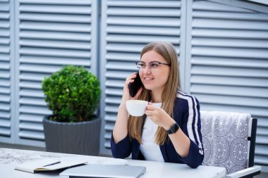 Dizüstü bilgisayarda çalışan ve telefonda konuşan güzel genç bir kadın kafede otururken gülümsüyor. Genç bir kadın iş için laptop kullanıyor. Açık bir kafede dizüstü bilgisayarda çalışan serbest çalışan bir kadın.