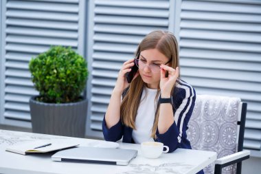 Dizüstü bilgisayarda çalışan ve telefonda konuşan güzel genç bir kadın kafede otururken gülümsüyor. Genç bir kadın iş için laptop kullanıyor. Açık bir kafede dizüstü bilgisayarda çalışan serbest çalışan bir kadın.