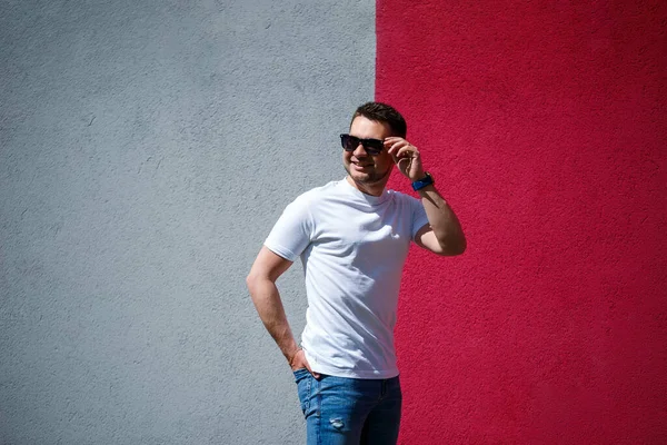 Beyaz tişört giymiş, gri ve kırmızı duvar arkasında duran yakışıklı bir adamın portresi. Şehir tarzı kıyafetler, modern moda imajı. Erkek modası