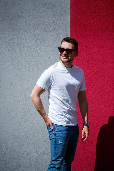 Tarz sahibi genç bir adamın portresi, beyaz bir tişört giymiş gri ve kırmızı bir duvar arkasında duran bir adam. Şehir tarzı kıyafetler, modern moda imajı. Erkek modası