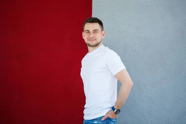 Stylish young man, a man dressed in a white blank t-shirt standing on a gray and red wall background. Urban style of clothes, modern fashionable image. Men\'s fashion