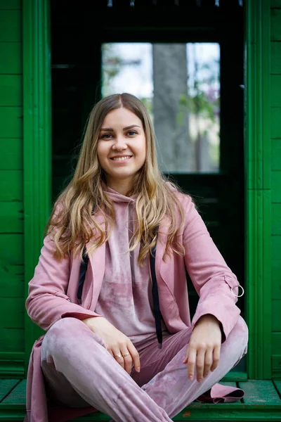 Avrupa görünümlü sarı saçlı şık genç kız merdivenlerde oturuyor, pembe bir takım elbise giyiyor, modern kadın modası, gündelik kıyafetler.