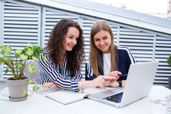 İki başarılı genç iş kadını yöneticisi bir dizüstü bilgisayar ve bir defterle bir masada oturmuş yeni bir geliştirme projesi üzerinde çalışıyor, bayan öğrenciler bilgisayarlarının çalışması üzerine bir rapor yazıyorlar.