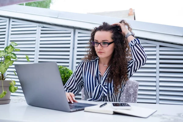 Başarılı genç bayan girişimci gülümseyerek bir bilgisayar ekranına bakıyor, açık kafe terasında dizüstü bilgisayar kullanarak çalışıyor.