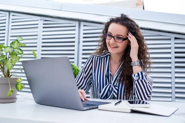 Başarılı genç bayan girişimci gülümseyerek bir bilgisayar ekranına bakıyor, açık kafe terasında dizüstü bilgisayar kullanarak çalışıyor.