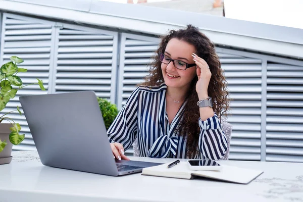 Başarılı genç bayan girişimci gülümseyerek bir bilgisayar ekranına bakıyor, açık kafe terasında dizüstü bilgisayar kullanarak çalışıyor.