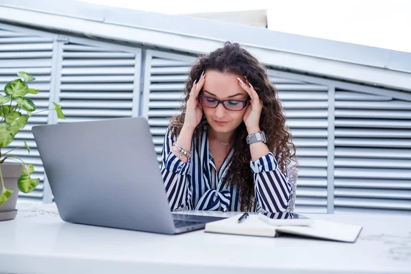 Moda kıyafetleri içinde serbest çalışan üzgün bir kadın, dizüstü bilgisayarla çalışıyor ve notlar alıyor, gülümsüyor ve dışarıdaki kafeye bakıyor. İş sorunları ve baş ağrısı