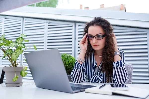 Moda kıyafetleri içinde serbest çalışan üzgün bir kadın, dizüstü bilgisayarla çalışıyor ve notlar alıyor, gülümsüyor ve dışarıdaki kafeye bakıyor. İş sorunları ve baş ağrısı