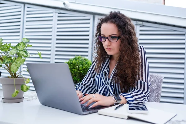 Moda modası geçmiş serbest çalışan, dizüstü bilgisayarla çalışan, notlar alan, gülümseyen ve açık hava teraslarına bakan düşünceli bir kadın.