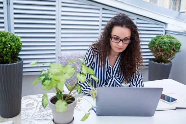 Moda modası geçmiş serbest çalışan, dizüstü bilgisayarla çalışan, notlar alan, gülümseyen ve açık hava teraslarına bakan düşünceli bir kadın.