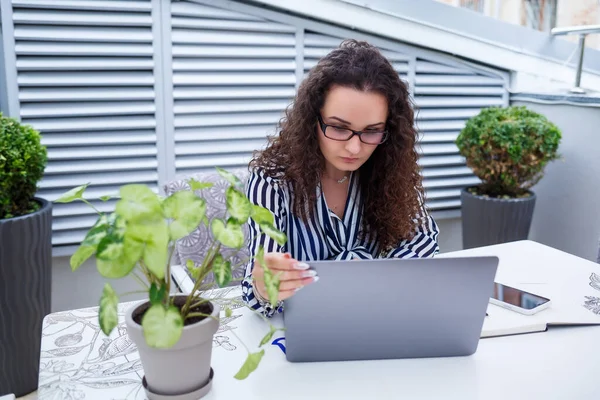 Moda modası geçmiş serbest çalışan, dizüstü bilgisayarla çalışan, notlar alan, gülümseyen ve açık hava teraslarına bakan düşünceli bir kadın.