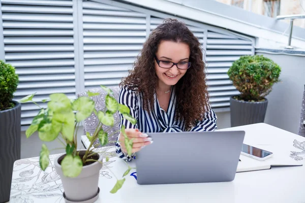 Moda modası geçmiş serbest çalışan, dizüstü bilgisayarla çalışan, notlar alan, gülümseyen ve açık hava teraslarına bakan düşünceli bir kadın.