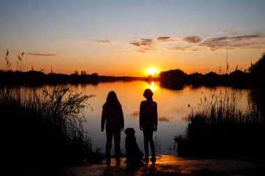 Gölde gün batımında köpekli kızların siluetleri, ağaçların arkasında güneş batıyor ve suda güzel yansımalar.
