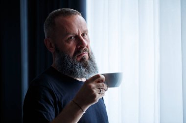 Sakallı yetişkin bir adam kahve içer ve pencereden dışarı bakar. Çalışma günü eğlence konsepti