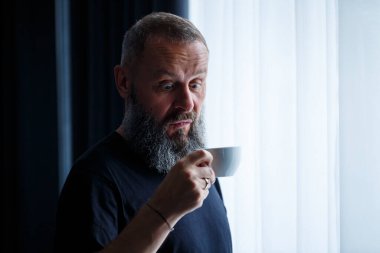 Sakallı yetişkin bir adam kahve içer ve pencereden dışarı bakar. Çalışma günü eğlence konsepti