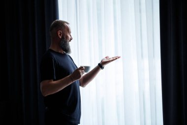 Sakallı yetişkin bir adam kahve içer ve pencereden dışarı bakar. Çalışma günü eğlence konsepti