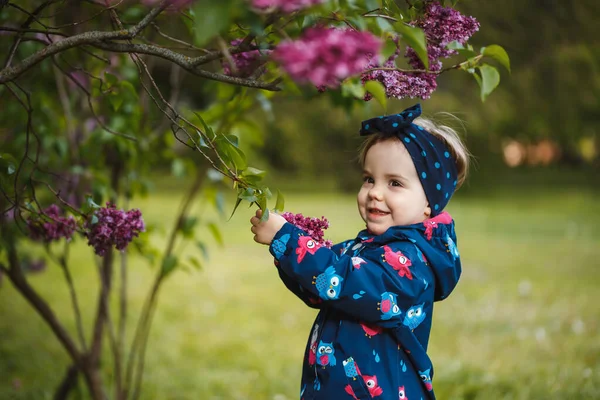 小さな女の子はライラックの緑豊かな茂みの近くに立って 彼女は笑顔と雪の紫色の花 — ストック写真