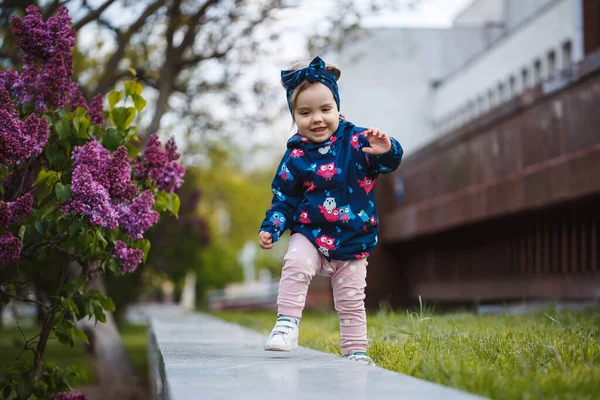 小さな女の子はライラックの緑豊かな茂みの近くに立って 彼女は笑顔と雪の紫色の花 — ストック写真