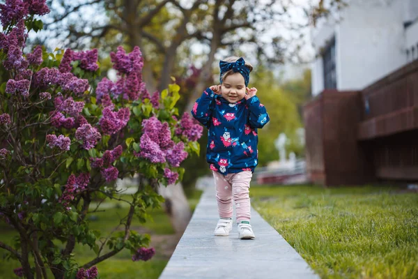 小さな女の子はライラックの緑豊かな茂みの近くに立って 彼女は笑顔と雪の紫色の花 — ストック写真