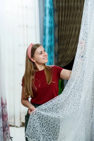 Young woman choosing fabric for new curtains in a store. Samples of the curtain hang on hangers on a rail in the store. Samples of textures of fabric, tulle and furniture upholstery.