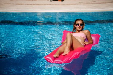 Seksi kadın model dinleniyor ve havuzda şiltede güneşleniyor. Pembe bikinili bir kadın şişme pembe bir yatakta yüzüyor. spf ve güneş kremi