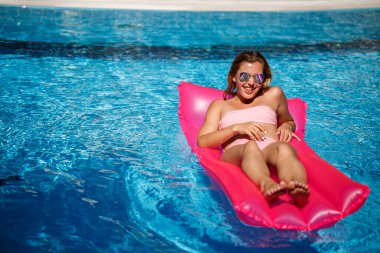 Seksi kadın model dinleniyor ve havuzda şiltede güneşleniyor. Pembe bikinili bir kadın şişme pembe bir yatakta yüzüyor. spf ve güneş kremi
