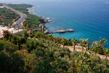 Boş bir asfalt yol güneşli bir yaz gününde nefes kesici kıyı şeridi boyunca esiyor. Berrak mavi gökyüzüne ve sakin Akdeniz 'e bakan kıyı yolunun muhteşem bir görüntüsü..