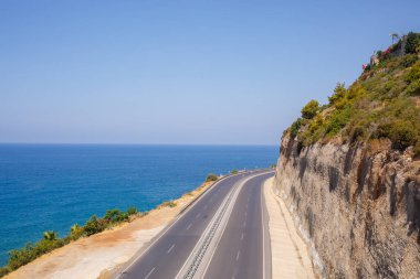 Boş bir asfalt yol güneşli bir yaz gününde nefes kesici kıyı şeridi boyunca esiyor. Berrak mavi gökyüzüne ve sakin Akdeniz 'e bakan kıyı yolunun muhteşem bir görüntüsü..