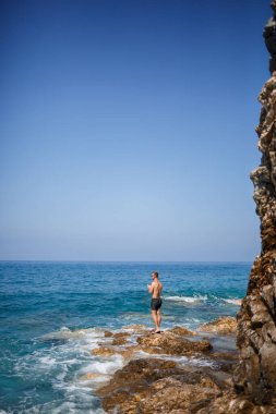 Genç bir adam açık Akdeniz manzaralı kayaların üzerinde duruyor. Sıcak bir yaz günü, güneşli bir günde bir adam deniz esintisine bakar.