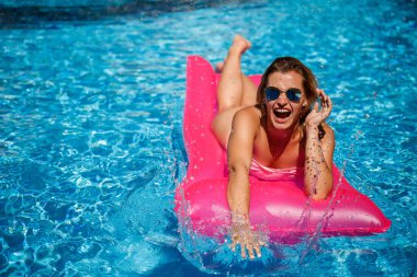Havuzdaki pembe şişme yatakta mayo giymiş seksi bir kadın yatıyor. Sıcak ve güneşli bir yaz gününde havuz kenarında rahatla. Tatil kavramı