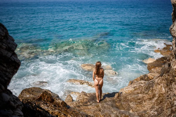 Sexy woman in full body swimsuit with long hair walks on large rocks on a rocky beach during a storm at sea. Back view. Woman swimwear island tropics landscape exotic walk