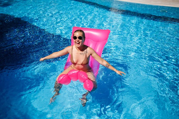戴着太阳镜的性感女人躺在游泳池的粉色床垫上晒太阳 穿着米黄色比基尼泳衣的年轻女子漂浮在充气粉色床垫上 — 图库照片