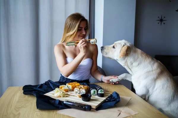 Beautiful blonde woman eating sushi close-up with her labrador dog, Smiling woman with lovely makeup holding a sushi roll with chopsticks. Healthy Japanese food. Diet concept