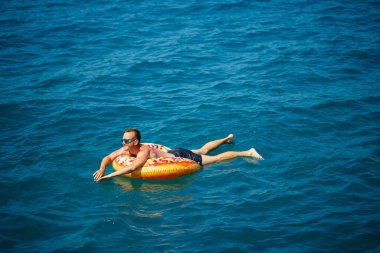 Genç bir adam mavi suyla denizde şişirilebilir bir halka üzerinde yüzüyor. Güneşli bir günde güzel bir tatil..