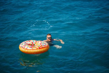 Genç adam güneşli bir günde denizde şişme bir yüzükte dinleniyor ve yüzüyor.