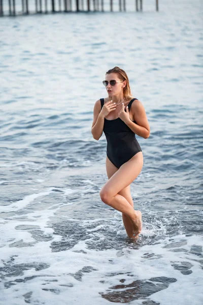 海によってビーチにポーズブロンドの髪と黒の水着で美しい女性の肖像画 ビーチを歩いている若い女性 — ストック写真
