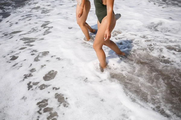 Two girls friends are walking along the sea sandy shore in swimsuits on a sunny warm day