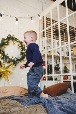 Neşeli bir çocuk, neşeli bir şekilde renkli, şenlikli süslemeler, parıldayan ışıklar ve sıcak, davetkar bir tatil atmosferiyle çevrili rahat bir yatakta zıplıyor.