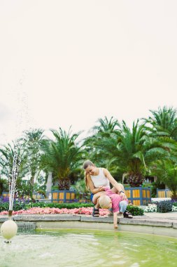 Canlı bir parkta yeşillik ve çiçeklerle, neşeli bir çocuk çeşmeden suya uzanırken, gülümseyen bir yetişkin aile ve arkadaşlarıyla keyifli anın tadını çıkarıyor.