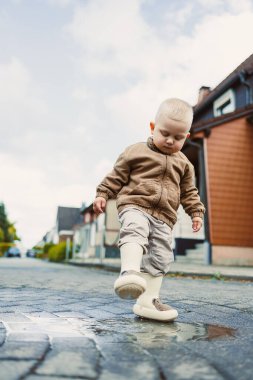 Genç bir çocuk ıslak bir caddedeki su birikintisine dikkatle adım atar, büyüleyici bir mahallede neşeli yağmurlu bir havada keşif yapmanın ve kahkaha atmanın saf mutluluğunu mutlulukla gösterir.