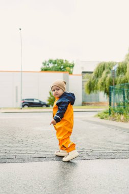 Canlı turuncu bir yağmurluk giymiş neşeli bir çocuk ve aynı şapkayı takmış ıslak bir sokakta neşeyle yürüyor, neşeli ruhlarını ve keyiflerini bu yağmurlu günde sergiliyor.