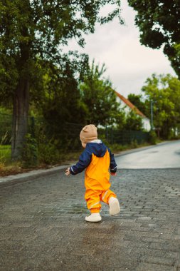 Neşeli bir çocuk, parlak ve canlı yağmur kıyafetleri içinde, neşe içinde boş bir kır yolunda koşmaktan zevk alıyor. Etrafı yemyeşil, karamsar bir gökyüzünün altında.