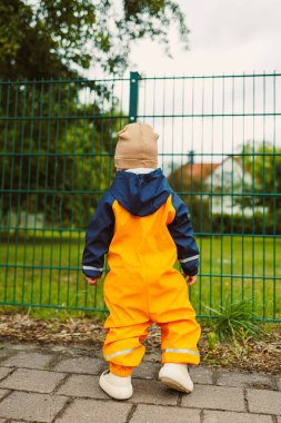 Parlak turuncu yağmurluk giymiş genç, maceraperest bir çocuk, bulutlu bir günde yeşil bir çitin kenarında dikilip olasılıklarla dolu açık bir alana bakıyor.