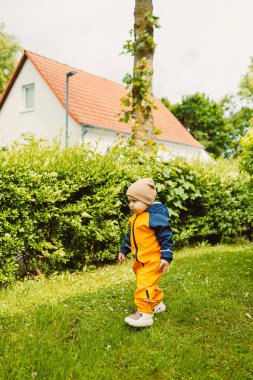 Neşeli ve canlı bir çocuk, parlak turuncu ve mavi yağmur giysileri içinde, neşeyle yürüyor yemyeşil bir bahçede, güzel güneşli bir günün tadını çıkarıyor.
