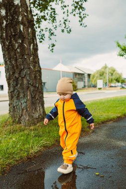 Parlak turuncu bir yağmurluk ve rahat bir bere giyen neşeli bir çocuk bulutlu bir gökyüzünün altındaki büyük su birikintilerine neşeyle sıçrıyor. Açık hava maceralarının heyecanını kucaklıyor.