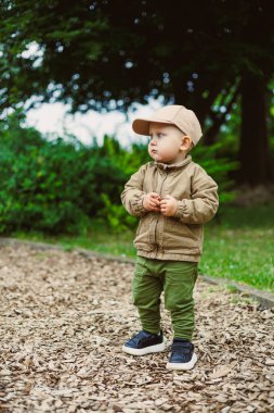 Şirin bir çocuk, neşeli bir şekilde parkta yumuşak odun parçalarının içinde duruyor, modaya uygun yeşil bir kıyafet ve aynı kepten giyinmiş, doğanın güzelliğini keşfetmeye hevesli.