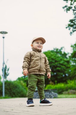Neşeli ve neşeli bir çocuk, neşeli bir şekilde dışarıda şık bir şekilde giyinmiş, neşe ve çocukluk masumiyetini çağrıştıran canlı yeşilliklerle çevrili duruyor.