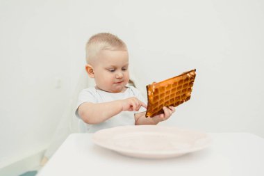 Meraklı küçük bir çocuk, minimalist beyaz bir masaya yerleştirilmiş parlak çikolatayı dikkatle inceliyor, çocukluk keşiflerinin masumiyetini, neşesini ve mucizesini güzelce yakalıyor.